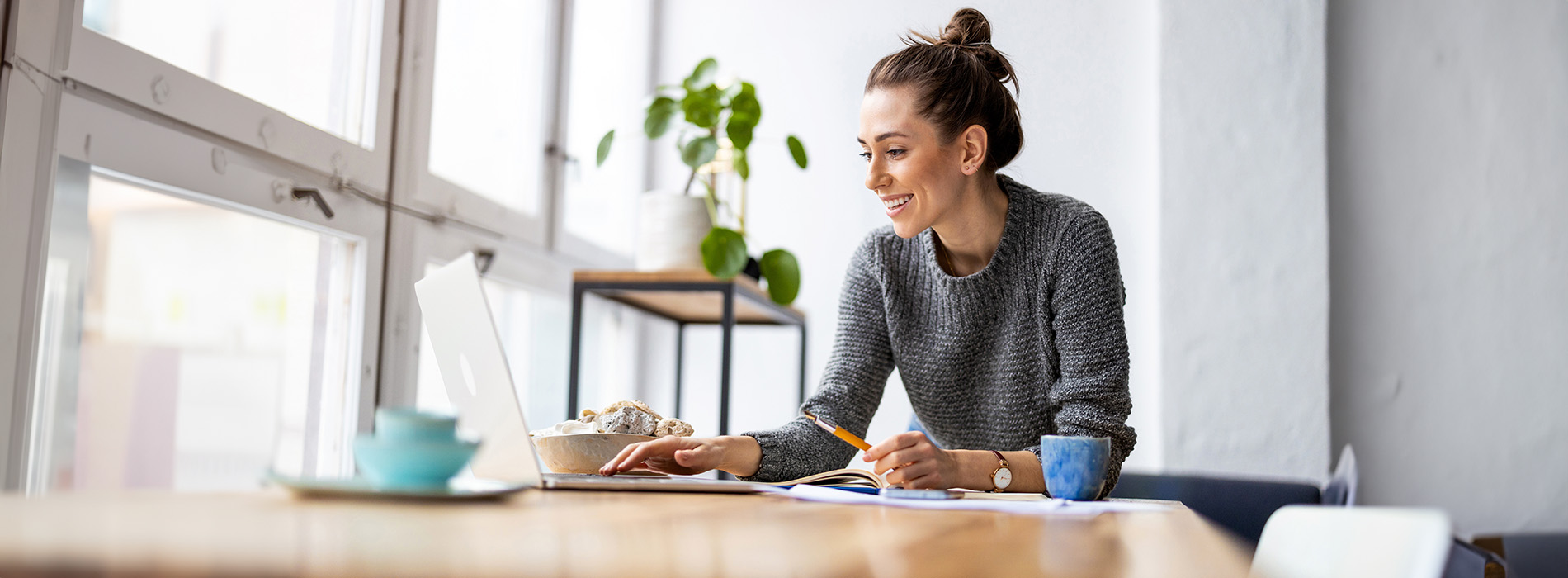 A woman sitting at a desk with a laptop, looking out a window, smiling, wearing a dark sweater and a necklace, with a potted plant nearby.